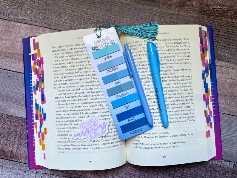 May include: A rectangular bookmark with a teal tassel, featuring a General Annotation Key with color-coded labels for Quotes, Plot Points, Love, Characters, Sad, Angry, and Additional. A blue pen rests on the bookmark, which lies on an open book.