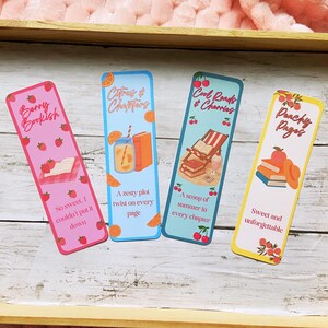 May include: Four colorful bookmarks with fruit and book-themed designs. The bookmarks feature text like "Berry Bookish" and "Peachy Pages." Each bookmark has a unique design, including strawberries, oranges, and cherries. They are displayed on a wooden tray.