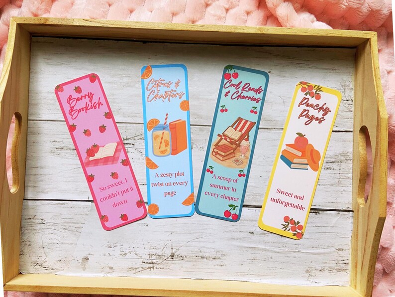 May include: Four colorful bookmarks with fruit and book-themed designs. The bookmarks feature text like "Berry Bookish" and "Peachy Pages." Each bookmark has a unique design, including strawberries, oranges, and cherries. They are displayed on a wooden tray.