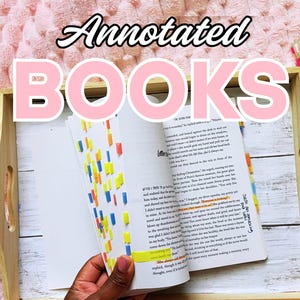 Könnte beinhalten: Ein offenes Buch mit Anmerkungen, farbigen Haftnotizen und markiertem Text liegt auf einem Holztablett. Der Text "Annotated BOOKS" ist in Rosa geschrieben. Der Hintergrund ist eine weiche, strukturierte rosa Oberfläche.
