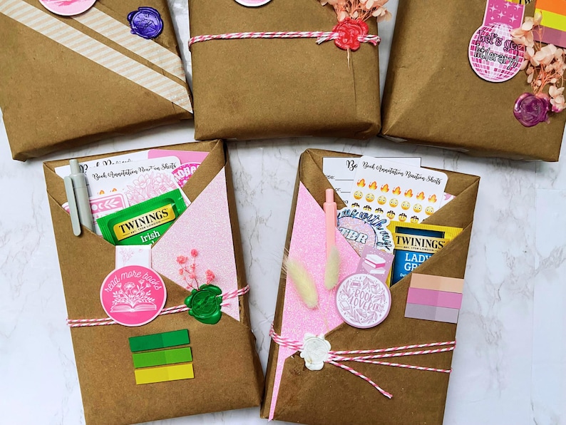 以下が含まれることがあります： ピンクと白の紐で結ばれた茶色の紙のパッケージ。ワックススタンプとステッカーで封印されています。一部のパッケージにはピンクのグリッターペーパーがあり、お茶、ペン、ステッカーが含まれています。「read more books」と「let's get literary」と書かれたステッカーで飾られています。