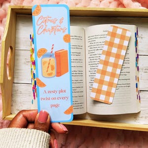 May include: A light blue bookmark with the text "Citrus & Chapters" and a zesty orange theme. The bookmark features a drink, orange slices, and a book illustration. An open book with a gingham bookmark rests on a wooden tray.