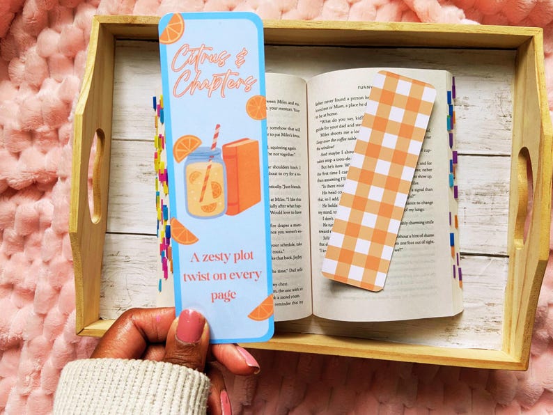 May include: A light blue bookmark with the text "Citrus & Chapters" and a zesty orange theme. The bookmark features a drink, orange slices, and a book illustration. An open book with a gingham bookmark rests on a wooden tray.
