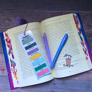May include: An open book with colorful page tabs, a purple tassel bookmark featuring a Fantasy Annotation Key, a light purple pen, a blue pen, and a sticker depicting an iced coffee with the text "coffee spicy books spicy" on the pages.