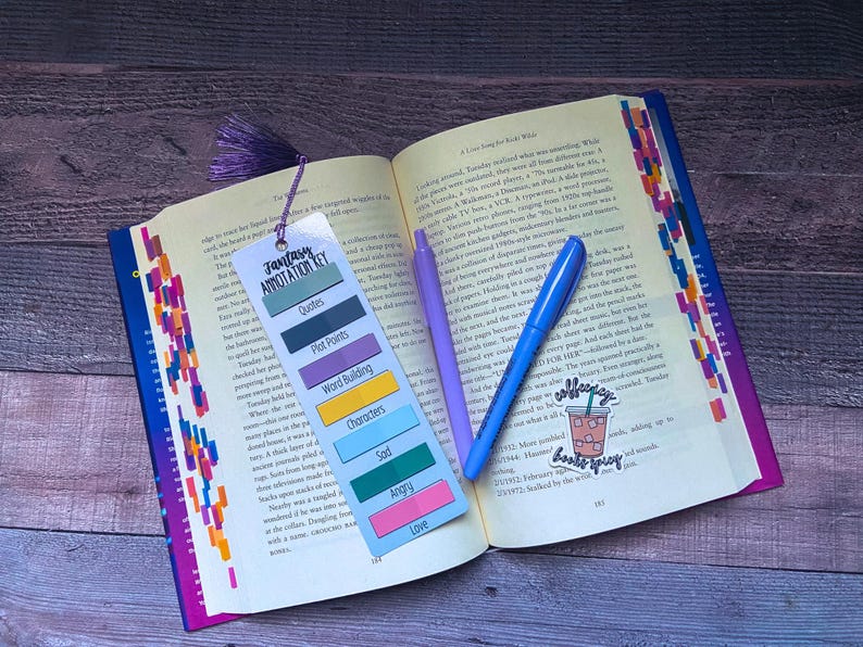 May include: An open book with colorful page tabs, a purple tassel bookmark featuring a Fantasy Annotation Key, a light purple pen, a blue pen, and a sticker depicting an iced coffee with the text "coffee spicy books spicy" on the pages.