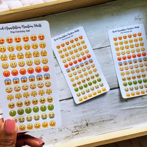 May include: Three sticker sheets with emoji faces in various expressions, including happy, sad, and angry. The stickers are arranged on white rectangular sheets with the text "Book Annotation Reaction Sheets The Fantasy Set". The sheets are on a wooden tray.
