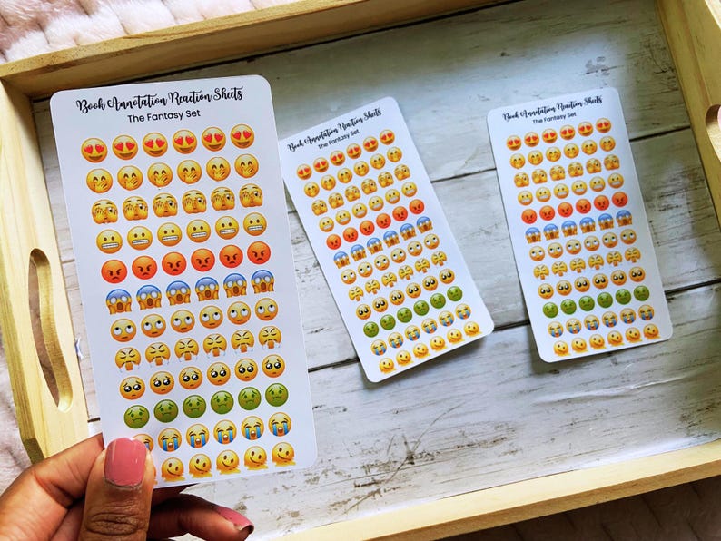 May include: Three sticker sheets with emoji faces in various expressions, including happy, sad, and angry. The stickers are arranged on white rectangular sheets with the text "Book Annotation Reaction Sheets The Fantasy Set". The sheets are on a wooden tray.
