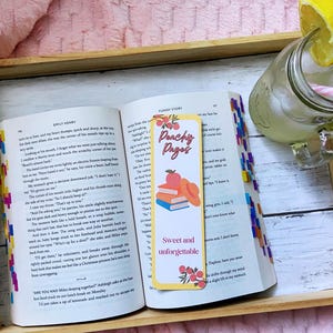 May include: An open book with colorful page markers and a bookmark that reads "Peachy Pages". A glass of lemonade with a lemon slice and a pink straw sits nearby on a wooden tray.