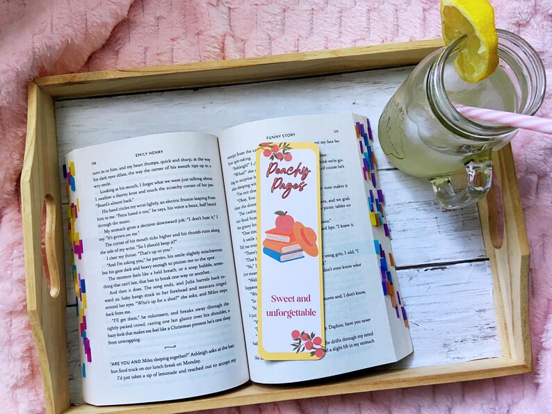 May include: An open book with colorful page markers and a bookmark that reads "Peachy Pages". A glass of lemonade with a lemon slice and a pink straw sits nearby on a wooden tray.