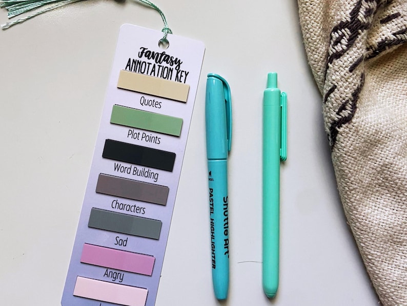 May include: A bookmark with the text "Fantasy Annotation Key" and color-coded sections for annotation. A teal highlighter and a mint green pen are next to the bookmark. A beige fabric with a dark brown pattern is in the background.