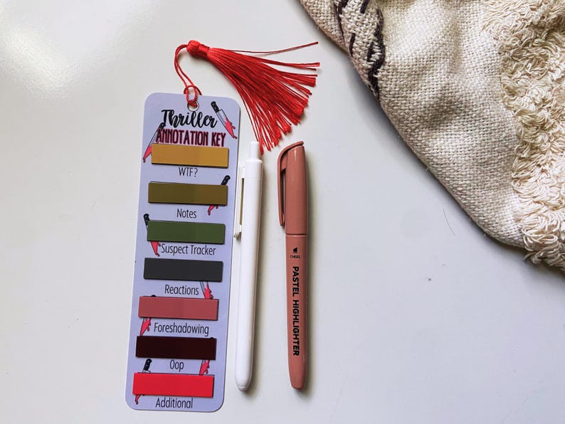 May include: A bookmark with a red tassel, labeled "Thriller Annotation Key," with color-coded sections for notes. A white pen and a pastel highlighter are next to the bookmark. The bookmark is designed for annotating thriller books.