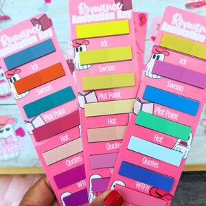 May include: Pink bookmark-style annotation keys with colorful labels for romance novels. Each key features text like "Ick," "Swoon," and "Plot Point," with a ghost cowboy graphic. The text "Romance Annotation Key" is at the top.
