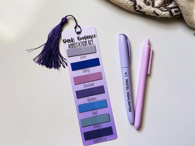 May include: A bookmark with a purple tassel, labeled "Dark Romance Annotation Key," features color-coded sections for "Love," "Spicy," and more. A pastel highlighter and a pink pen are also present.