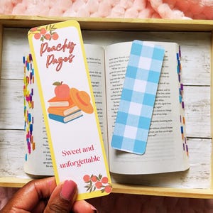 May include: A wooden tray holds an open book with two bookmarks. One bookmark is yellow with the text "Peachy Pages" and a peach and book illustration. The other is a blue and white gingham pattern.
