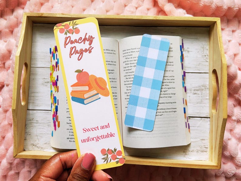 May include: A wooden tray holds an open book with two bookmarks. One bookmark is yellow with the text "Peachy Pages" and a peach and book illustration. The other is a blue and white gingham pattern.