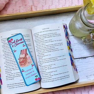 May include: An open book with a blue bookmark that reads "Cool Reads & Cherries" and "A scoop of summer in every chapter." A glass of lemonade with a lemon slice and a pink straw sits on a wooden tray.