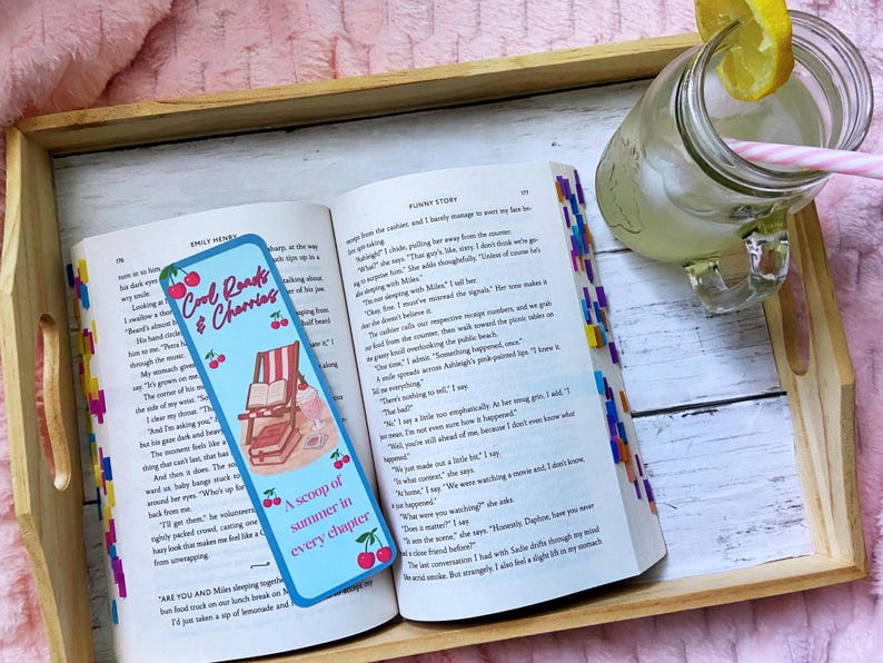 May include: An open book with a blue bookmark that reads "Cool Reads & Cherries" and "A scoop of summer in every chapter." A glass of lemonade with a lemon slice and a pink straw sits on a wooden tray.