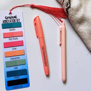 May include: A bookmark with a General Annotation Key, featuring color-coded labels for Quotes, Plot Points, Love, Characters, Sad, Angry, and Additional. Includes a coral marker and a light pink pen. A red tassel hangs from the bookmark.