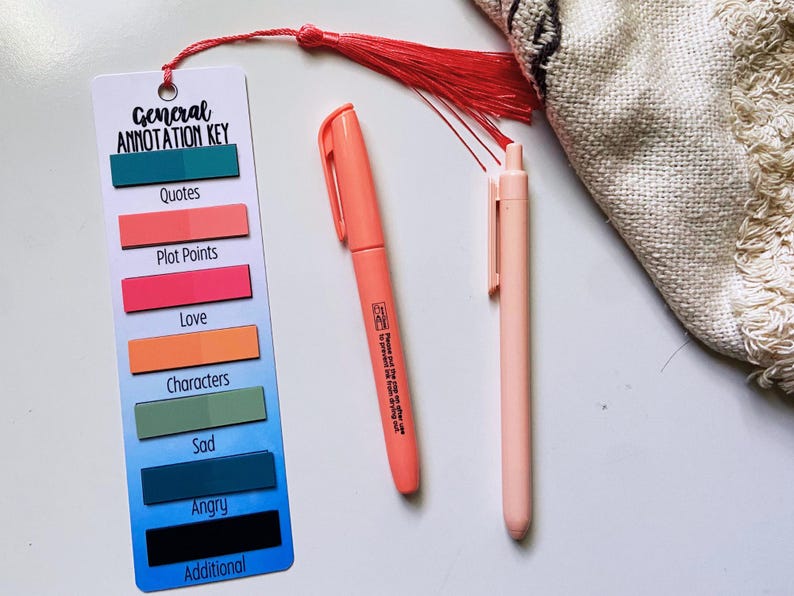 May include: A bookmark with a General Annotation Key, featuring color-coded labels for Quotes, Plot Points, Love, Characters, Sad, Angry, and Additional. Includes a coral marker and a light pink pen. A red tassel hangs from the bookmark.