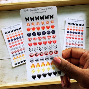 May include: A hand holds sticker sheets with various emoji designs, including butterflies, hearts, mushrooms, and musical notes. The text "Book Annotation Reaction Sheets" and "The After Dark Set" is visible. The stickers are arranged on a wooden tray.
