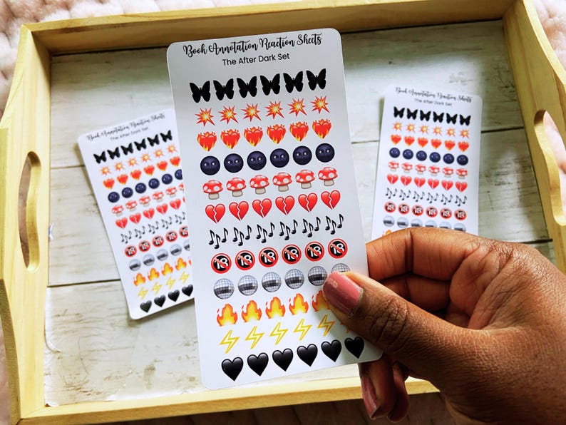 May include: A hand holds sticker sheets with various emoji designs, including butterflies, hearts, mushrooms, and musical notes. The text "Book Annotation Reaction Sheets" and "The After Dark Set" is visible. The stickers are arranged on a wooden tray.