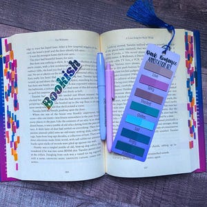 May include: An open book with a colorful bookmark featuring the text Dark Romance Annotation Key, listing categories like Love, Spicy, Shocked, Quotes, Sad, Angry, and Additional. A Bookish sticker and two light-colored pens rest on the pages.