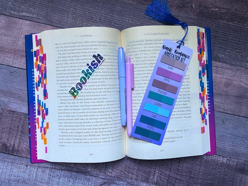 May include: An open book with a colorful bookmark featuring the text Dark Romance Annotation Key, listing categories like Love, Spicy, Shocked, Quotes, Sad, Angry, and Additional. A Bookish sticker and two light-colored pens rest on the pages.
