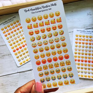 May include: Three sticker sheets with emoji faces in various expressions, including smiling, laughing, and crying. The sheets are labeled "Book Annotation Reaction Sheets" and "The Contemporary Set". The stickers are arranged on a wooden tray.