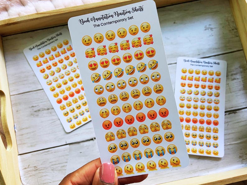 May include: Three sticker sheets with emoji faces in various expressions, including smiling, laughing, and crying. The sheets are labeled "Book Annotation Reaction Sheets" and "The Contemporary Set". The stickers are arranged on a wooden tray.
