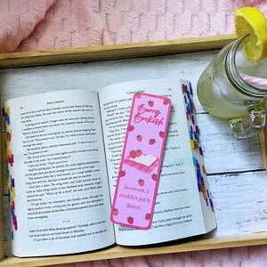 May include: An open book with a pink bookmark that reads "Berry Bookish" and "So sweet, I couldn't put it down." A glass of lemonade with a lemon slice and a pink straw sits next to the book on a wooden tray.