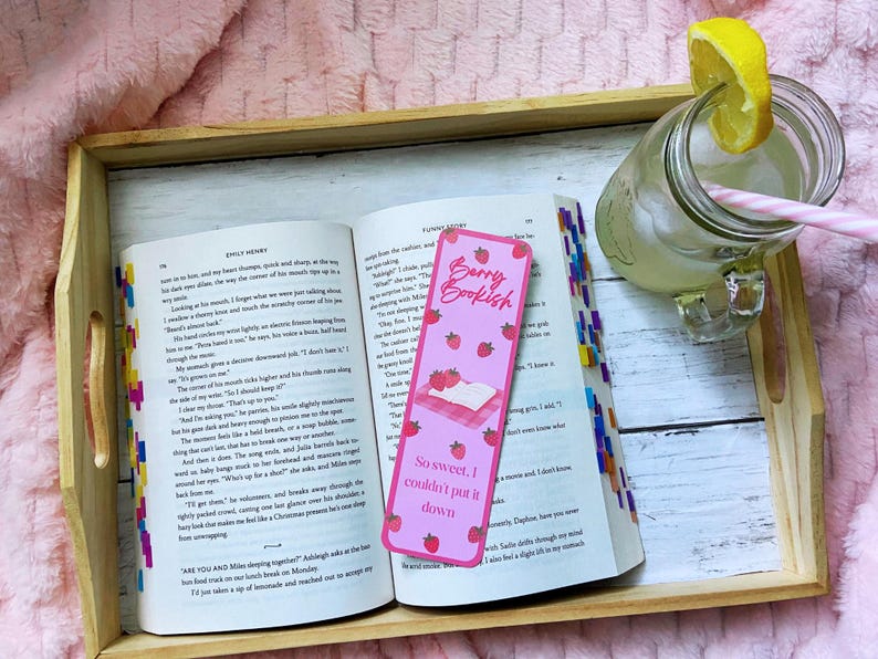 May include: An open book with a pink bookmark that reads "Berry Bookish" and "So sweet, I couldn't put it down." A glass of lemonade with a lemon slice and a pink straw sits next to the book on a wooden tray.