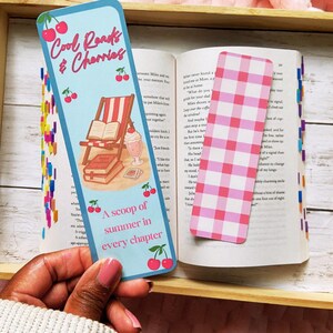 May include: A blue bookmark with the text "Cool Reads & Cherries" and "A scoop of summer in every chapter" is held over an open book. The bookmark features a cherry and beach scene illustration. A pink and white checkered bookmark is also visible.
