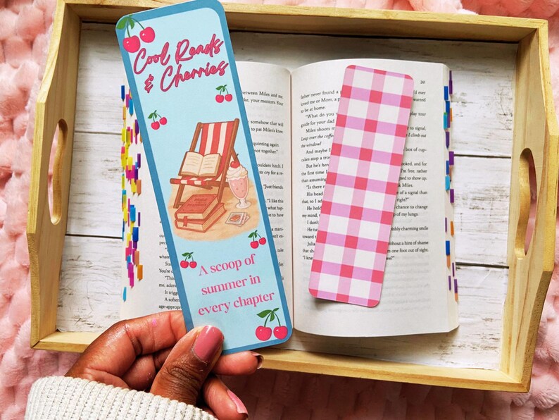 May include: A blue bookmark with the text "Cool Reads & Cherries" and "A scoop of summer in every chapter" is held over an open book. The bookmark features a cherry and beach scene illustration. A pink and white checkered bookmark is also visible.