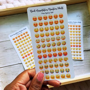 May include: Three sticker sheets with emoji faces in various expressions, including hearts for eyes, smiling, laughing, and crying. The text "Book Annotation Reaction Sheets" and "The Spicy Set" are printed on the stickers. The stickers are on a wooden tray.