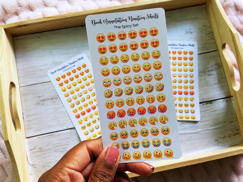 May include: Three sticker sheets with emoji faces in various expressions, including hearts for eyes, smiling, laughing, and crying. The text "Book Annotation Reaction Sheets" and "The Spicy Set" are printed on the stickers. The stickers are on a wooden tray.
