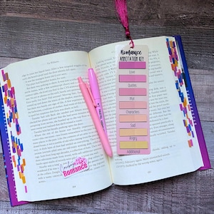May include: An open book with colorful page markers, a pink highlighter, and a vertical bookmark featuring a Romance Annotation Key. The bookmark lists categories including Love, Quotes, Plot, Characters, Sad, Angry, and Additional, each with a corresponding color block and a dark pink tassel at the top.