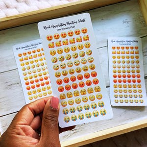 May include: Three sticker sheets with emoji faces, including hearts, smiling, and angry faces. The text "Book Annotation Reaction Sheets" and "The General Set" is printed on the stickers. The stickers are on a wooden tray.
