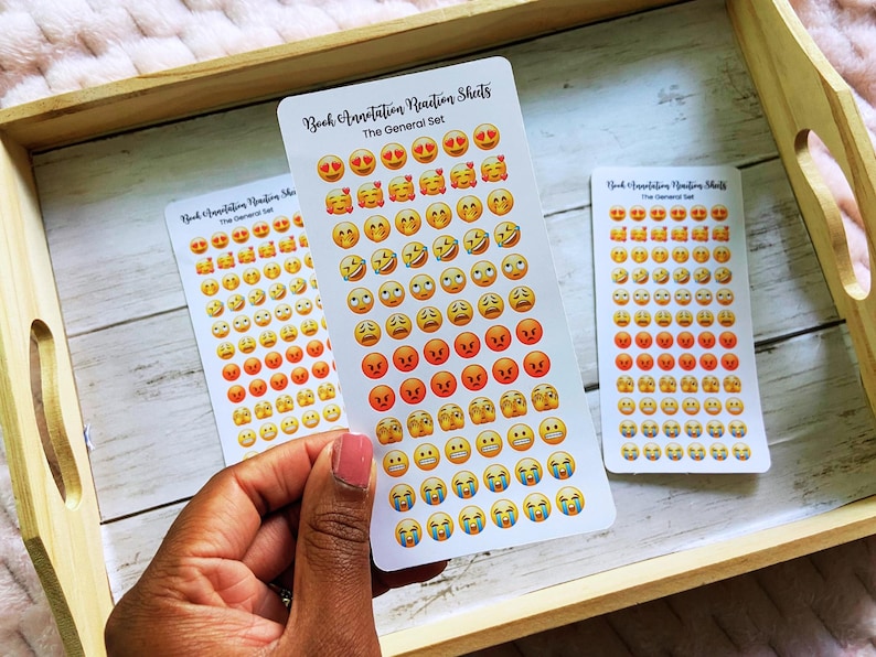 May include: Three sticker sheets with emoji faces, including hearts, smiling, and angry faces. The text "Book Annotation Reaction Sheets" and "The General Set" is printed on the stickers. The stickers are on a wooden tray.