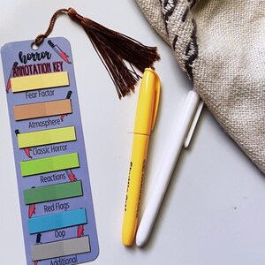 May include: A bookmark with the text "horror ANNOTATION KEY" and colorful sticky notes for categorizing horror elements. A yellow marker and a white pen are next to the bookmark. A brown tassel hangs from the top of the bookmark.