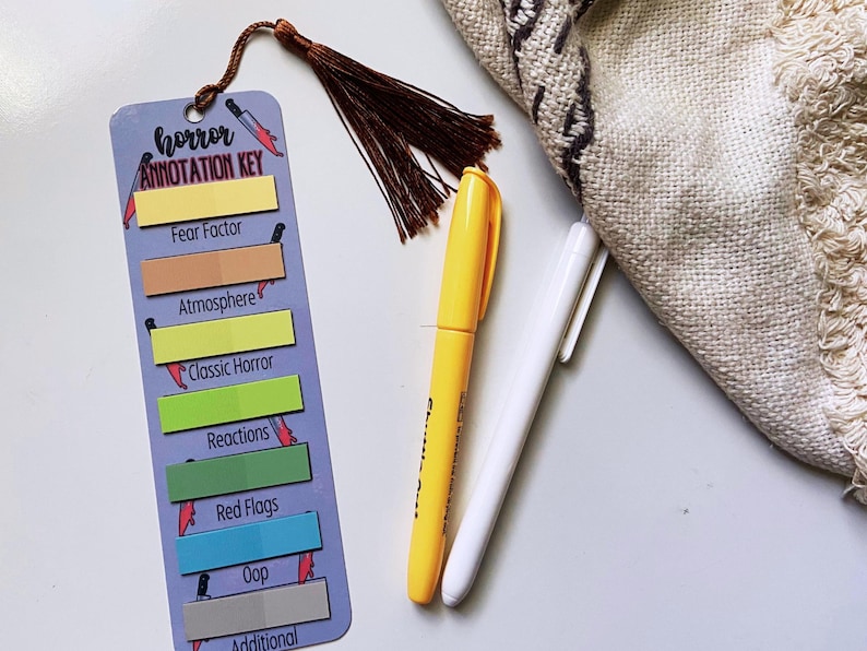 May include: A bookmark with the text "horror ANNOTATION KEY" and colorful sticky notes for categorizing horror elements. A yellow marker and a white pen are next to the bookmark. A brown tassel hangs from the top of the bookmark.