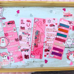 May include: A wooden tray displays pink bookmarks and stickers with a cowboy theme. The bookmarks feature phrases like "Cowboys don't cry" and "Save a horse ride a cowboy." Stickers include cowboy boots, hats, and ghost illustrations. Hearts and text accents complete the design.