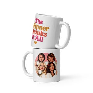 Puede incluir: Dos tazas de cerámica blanca apiladas verticalmente. La taza superior presenta el texto en rosa y naranja "The Winner Drinks It All" con un pequeño corazón naranja. La taza inferior muestra un gráfico circular de cuatro personas sosteniendo tazas blancas.