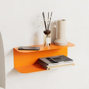 Può includere: Una mensola galleggiante in metallo arancione brillante a due livelli, montata su una parete bianca. Il ripiano superiore contiene uno smartphone, un diffusore in vetro e un vaso beige. Il ripiano inferiore mostra una pila di libri.