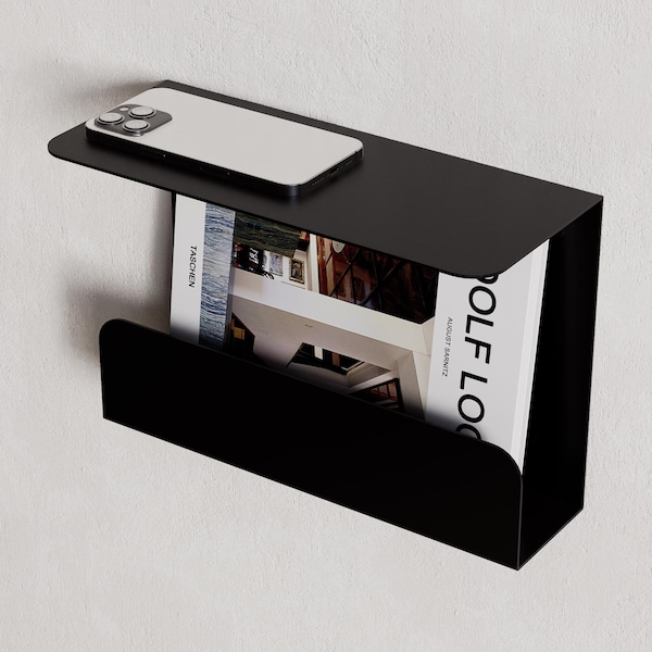 May include: A black metal bedside shelf mounted on a white wall. A smartphone rests on the top shelf, and a magazine is stored in the lower compartment. The magazine's title is partially visible: "GOLF LOG".