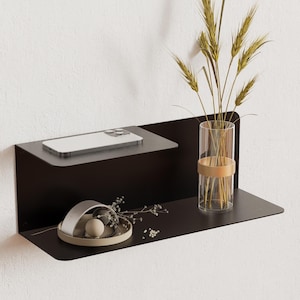 May include: A black metal floating shelf fixed to a white wall. The shelf has two tiers, with a smartphone on the upper tier and a glass vase with wheat stalks on the right. A small decorative dish is on the lower tier.