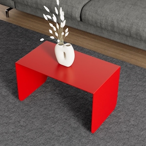 May include: A modern red coffee table with a minimalist design. The table is rectangular with a solid top and two side panels. A white vase with decorative white stems sits on top of the table. The table is on a gray rug.