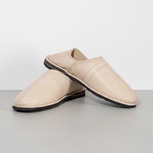 Puede incluir: Un par de zapatillas de cuero beige con suela negra. Las zapatillas se muestran sobre una superficie gris claro. Las zapatillas tienen un diseño sencillo con una costura cosida en la parte superior.
