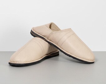 May include: A pair of beige leather slippers with a black sole. The slippers are shown on a light gray surface. The slippers have a simple design with a stitched seam across the top.