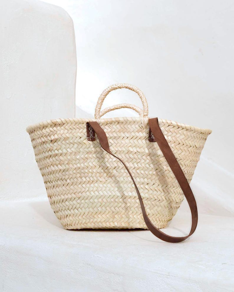 May include: A woven straw tote bag with two small handles and a long brown leather strap. The bag is a light beige color and has a rectangular shape with a wide opening. The bag is sitting on a white surface.