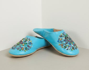 Puede incluir: Un par de zapatillas de cuero azul claro con intrincados diseños florales de cuentas de varios colores. Las zapatillas tienen una punta redondeada y una suela de color neutro. La imagen está tomada desde un ángulo ligeramente elevado, mostrando los detalles.